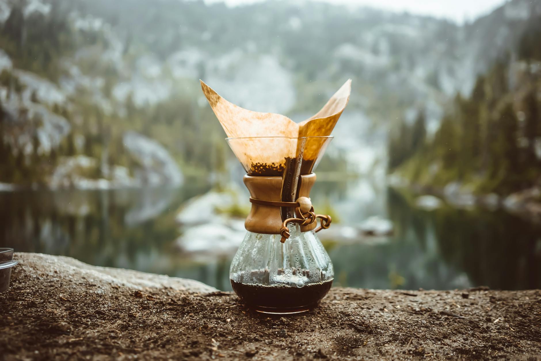 Chemex coffee pot brewing in a scenic mountain setting by a tranquil lake.